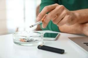 A person holds a cigarette over an ashtray; behind are a smartphone, vaping device, and a partially visible laptop on a white surface.