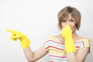 A person with short hair, wearing a striped shirt and yellow cleaning gloves, points to the side, covering their mouth, and looking surprised.