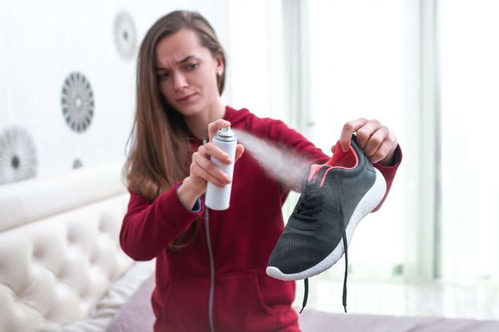 A person is spraying deodorizer on a black and red sneaker, with a concentrated look, indoors with light decor in the background.