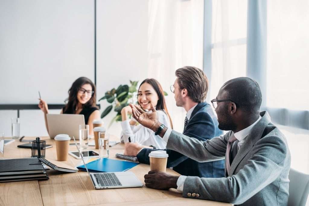 Four people engage in a discussion in a well-lit meeting room with a laptop, notebooks, and coffee cups on the table, exchanging ideas in a collaborative atmosphere.