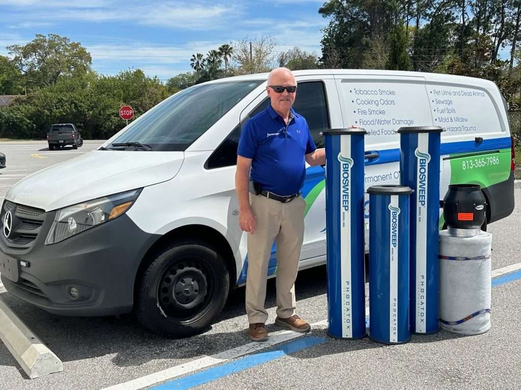 A person is standing next to a commercial vehicle with signage, holding equipment possibly related to odor elimination, in a sunny parking lot with trees.